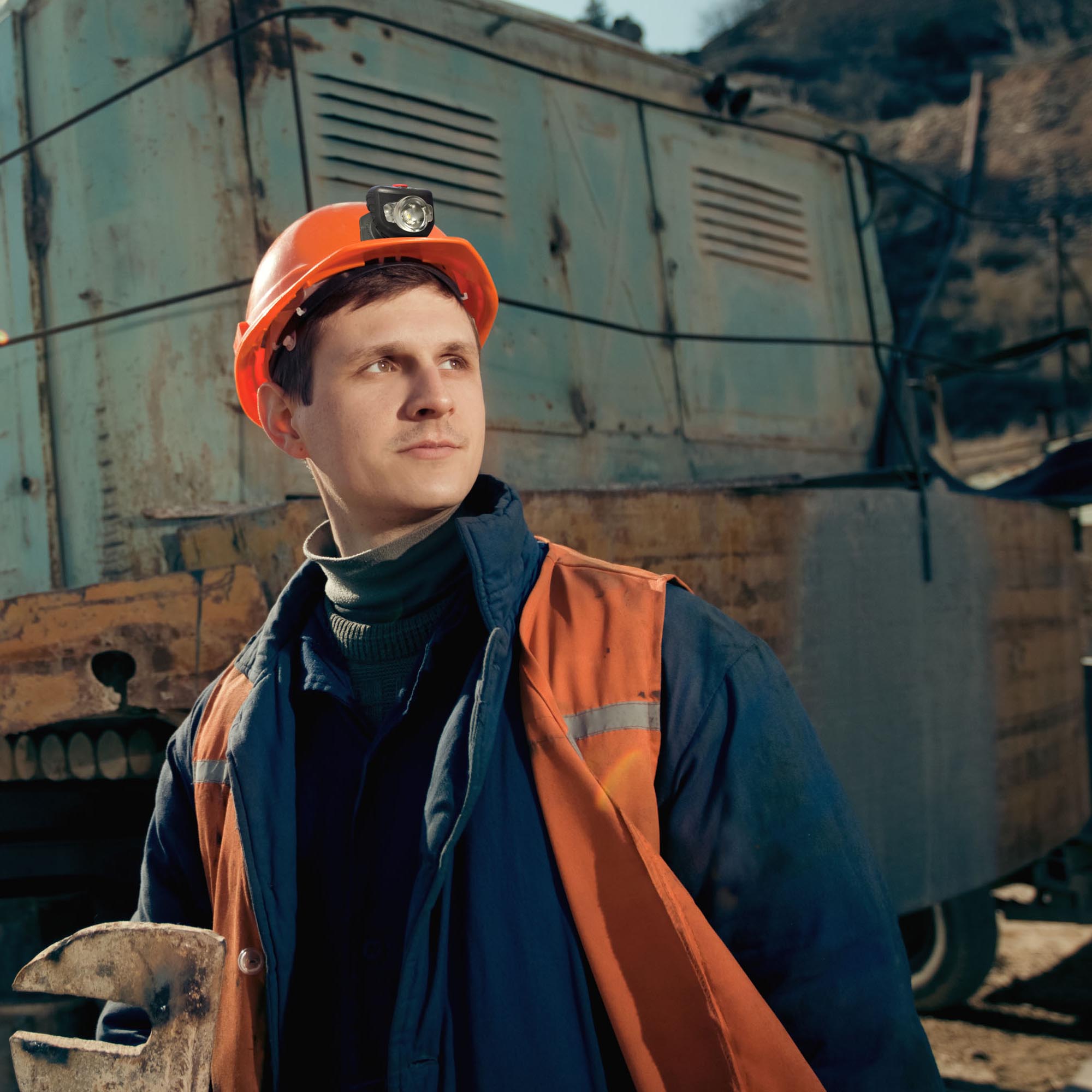 Person wearing an orange hard hat and safety vest in front of a large piece of mining equipment.